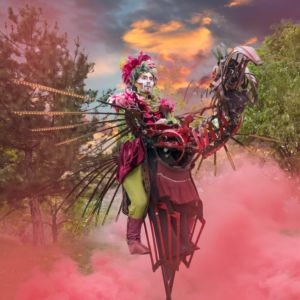 Un artiste en échasses, maquillé et costumé, chevauche une créature fantastique articulée. Des plumes, des lumières et des structures métalliques évoquent un oiseau mécanique. Une fumée rose entoure la scène, sous un ciel aux couleurs éclatées.