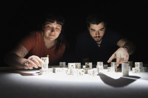 Une femme et un homme, concentrés, manipulent des petits cubes blancs éclairés par un faisceau de lumière dans l’obscurité. Leurs visages sont partiellement éclairés, créant une ambiance intime et immersive.